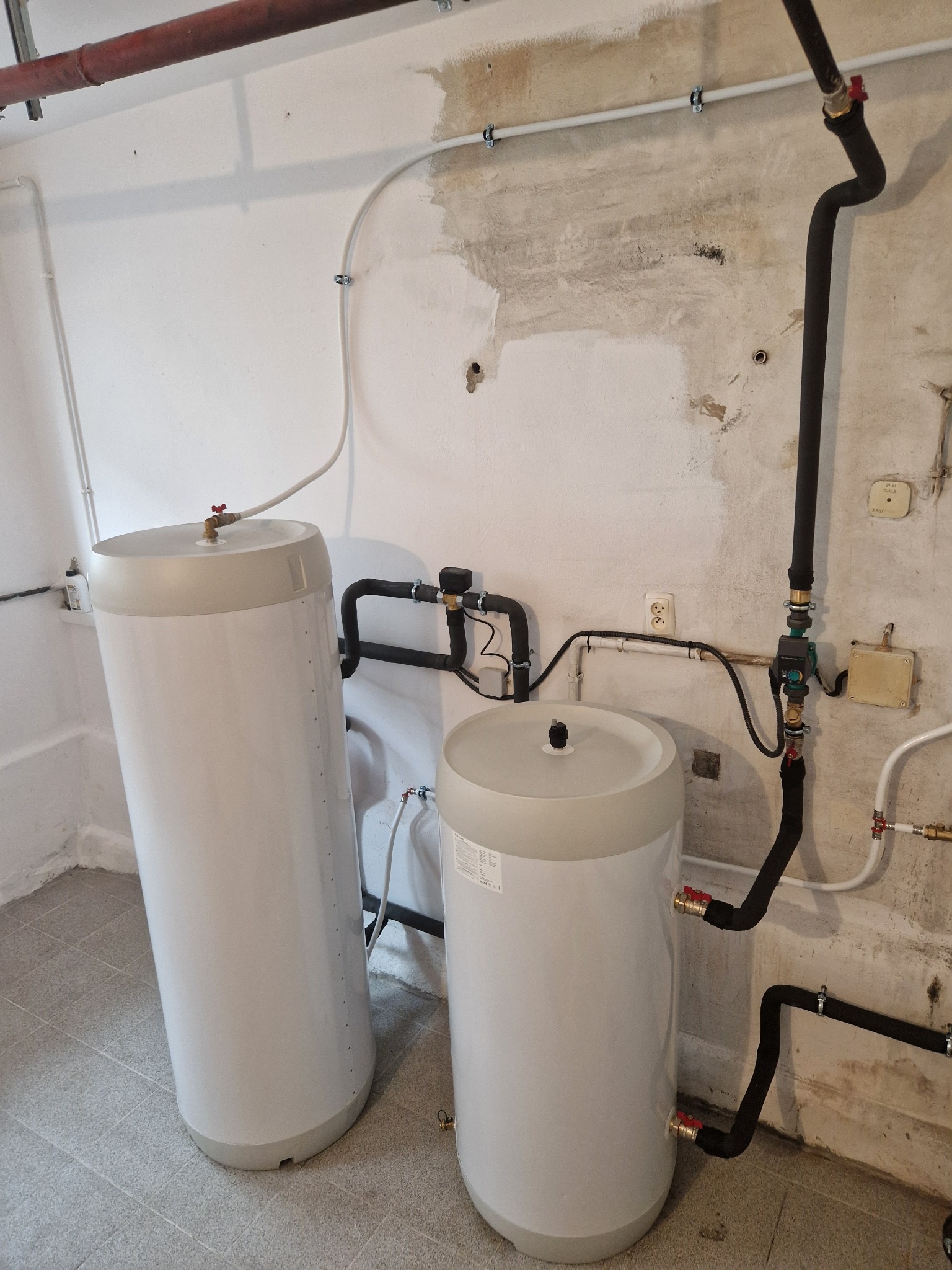 Dwa białe zbiorniki akumulacyjne ciepła z szarymi pokrywami, podłączone do instalacji rurowej w piwnicy, widoczne rury miedziane i czarne izolowane, ściana z widocznymi śladami napraw i instalacji...
