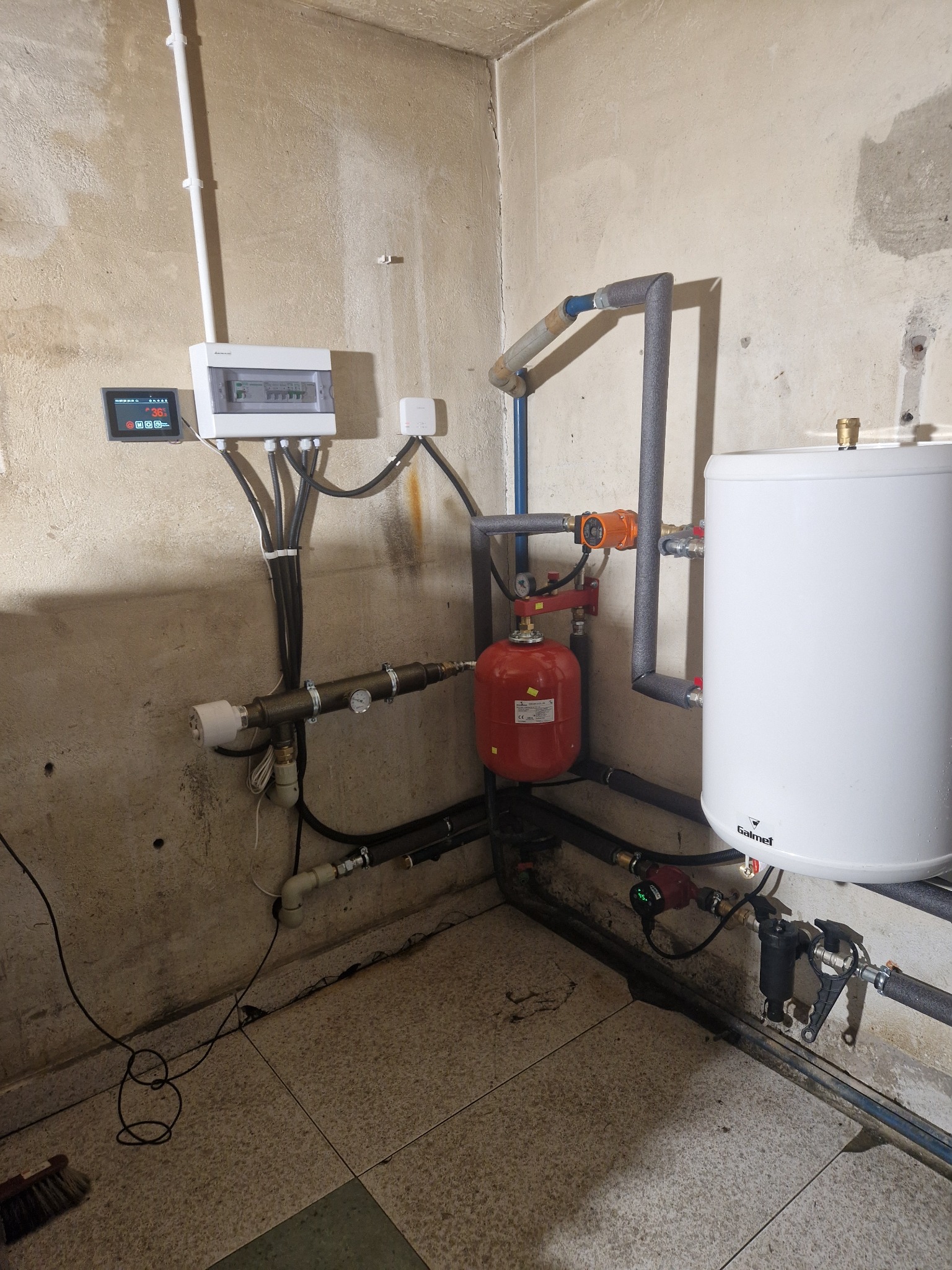 Instalacja kotłowni w piwnicy: biały zasobnik ciepłej wody Galmet, czerwony zbiornik wyrównawczy, orurowanie z izolacją, szafka elektryczna z wyświetlaczem, pompa obiegowa i filtr, na tle surowych...