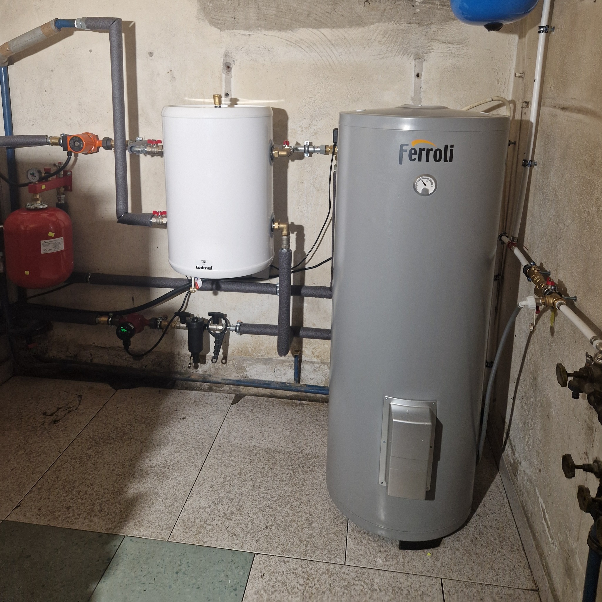 Dwa zbiorniki na wodę: biały Galmet i szary Ferroli, połączone instalacją grzewczą z rurami w izolacji, widoczne pompy obiegowe i naczynie przeponowe w kotłowni.