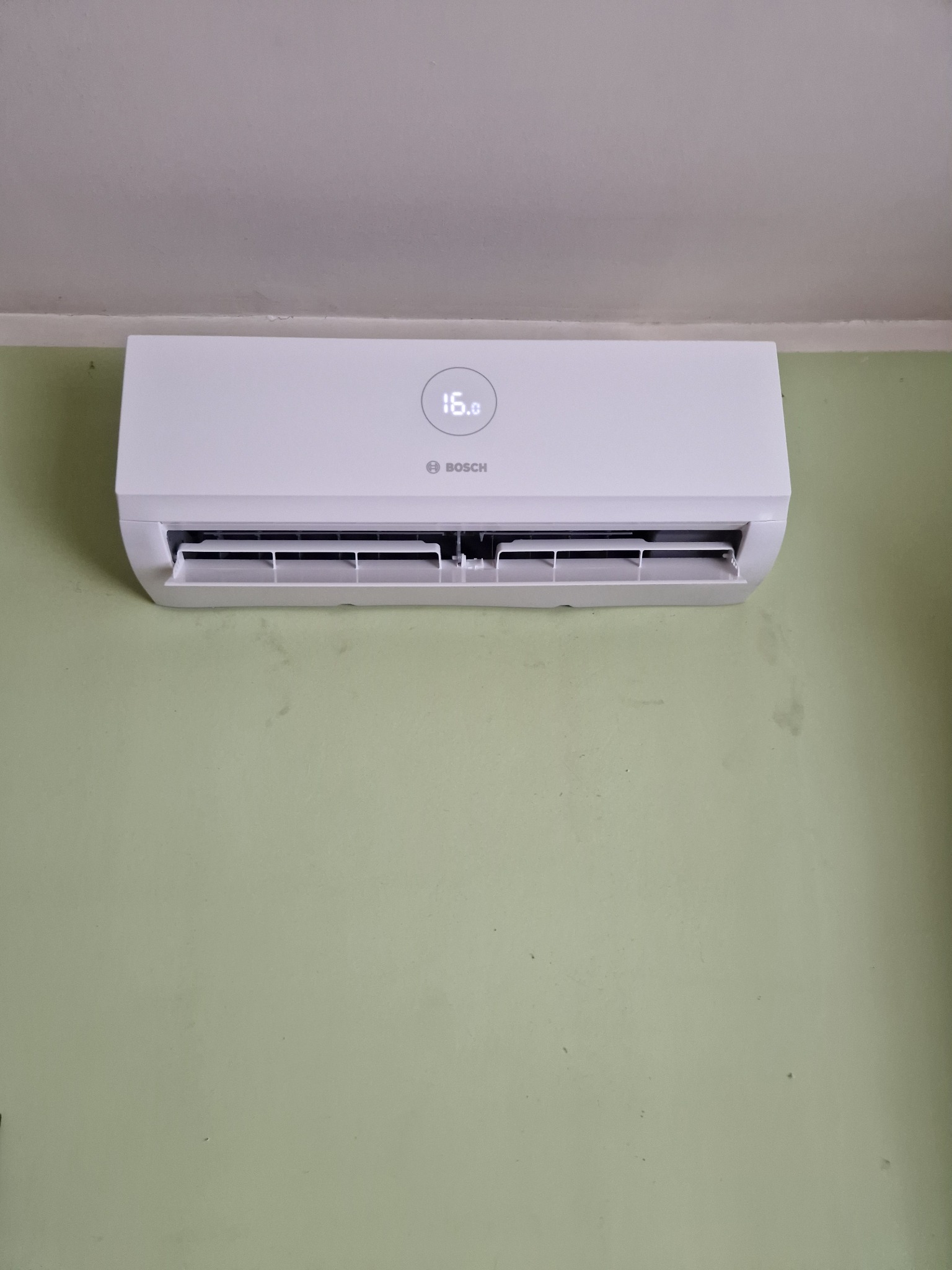 Biała jednostka klimatyzacyjna Bosch zamontowana na zielonej ścianie, wyświetlająca temperaturę 16 stopni Celsjusza.