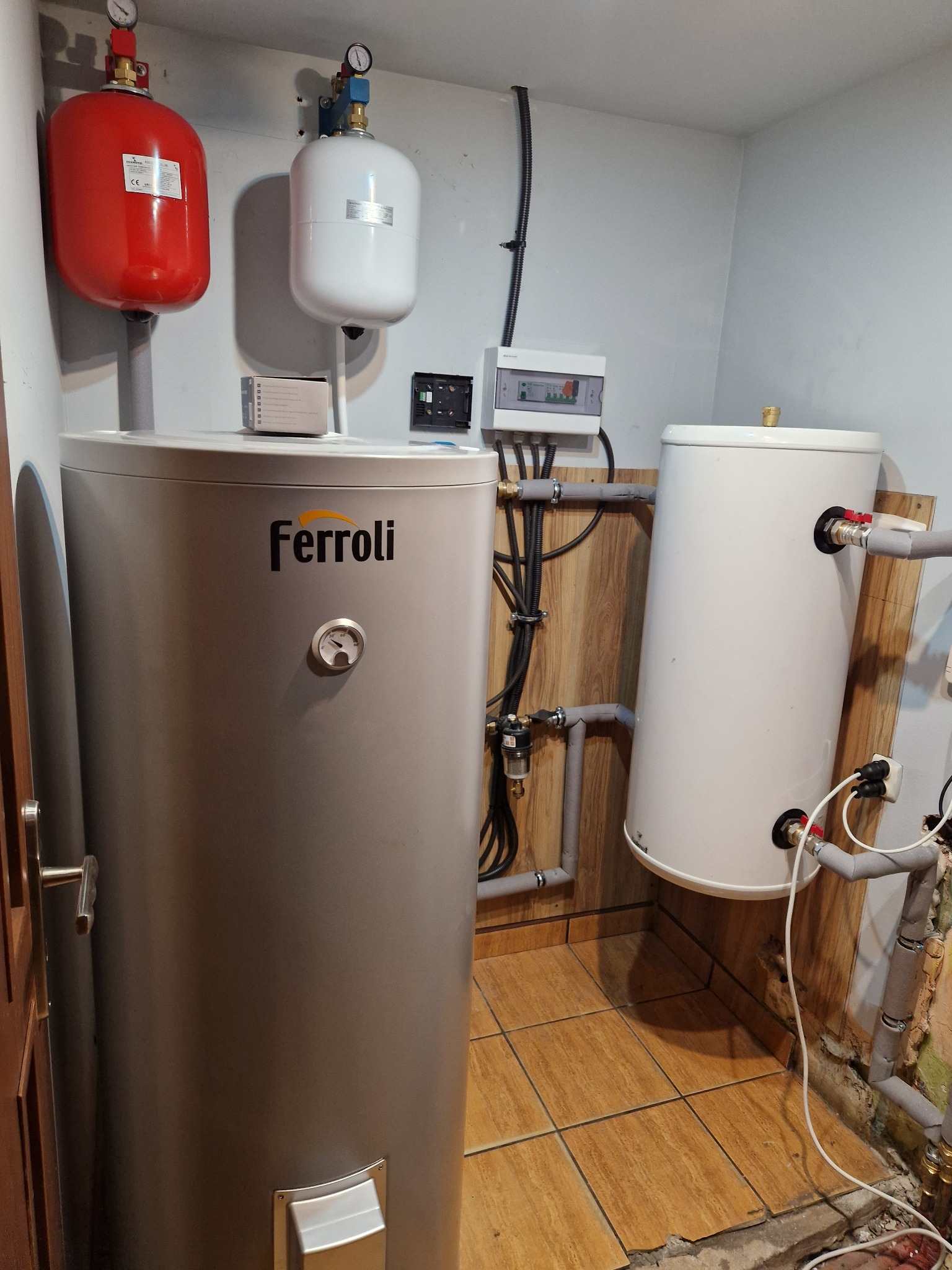 Instalacja grzewcza w kotłowni: srebrny zasobnik ciepłej wody marki Ferroli z termometrem, czerwony i biały zbiornik wyrównawczy, orurowanie, skrzynka elektryczna na ścianie, brązowe płytki podłogowe.