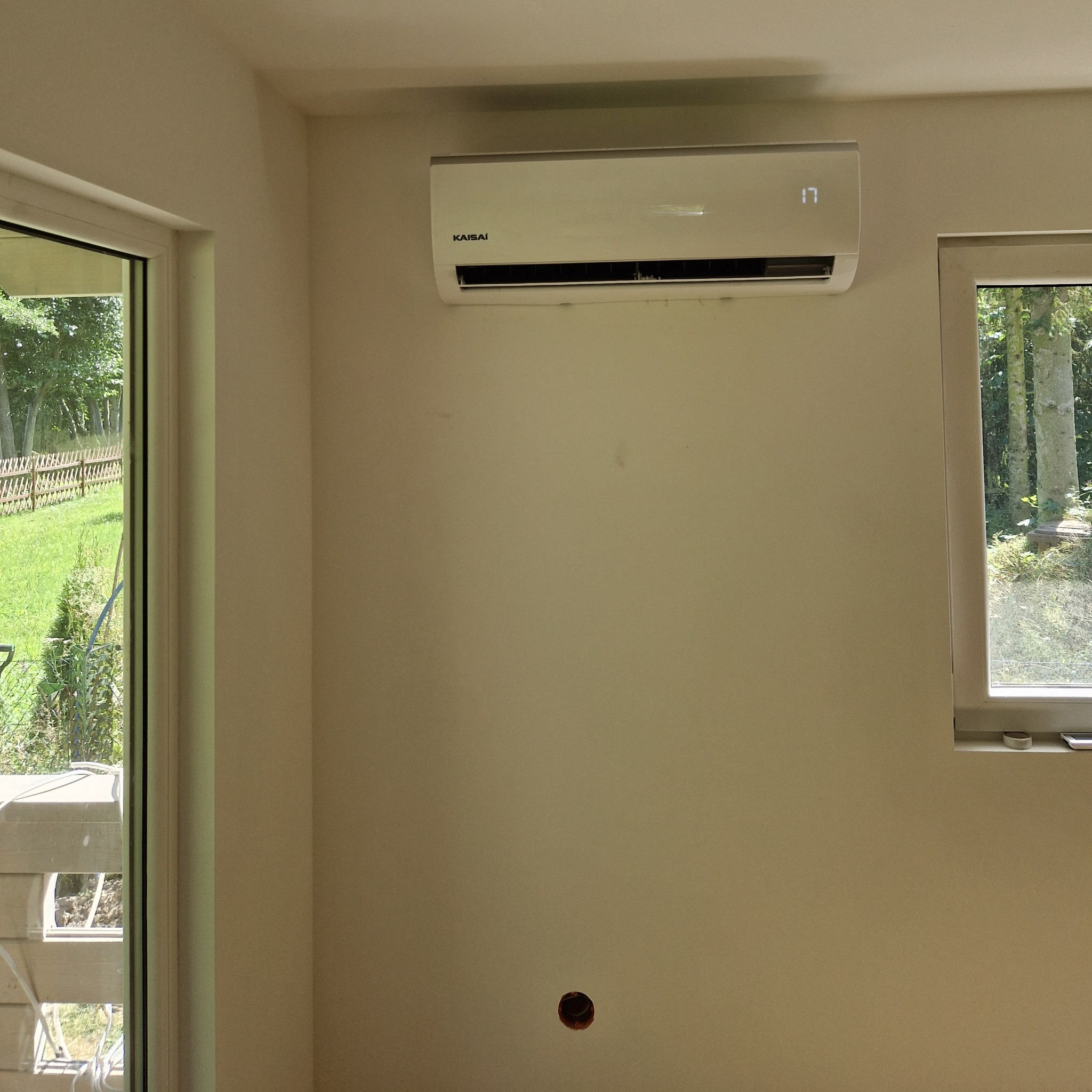 Wnętrze pokoju z zamontowanym klimatyzatorem Kaisai na ścianie, widoczne okno z widokiem na zieleń i otwór w ścianie pod instalację.