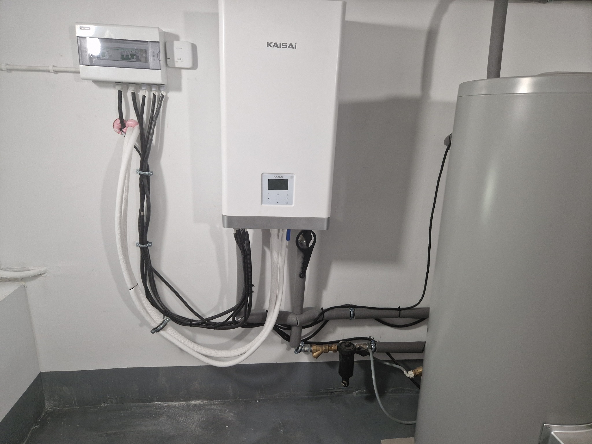 Biała jednostka wewnętrzna z wyświetlaczem marki Kaisai, podłączona do instalacji elektrycznej i zbiornika na wodę, widoczne rury i przewody, instalacja w pomieszczeniu technicznym.