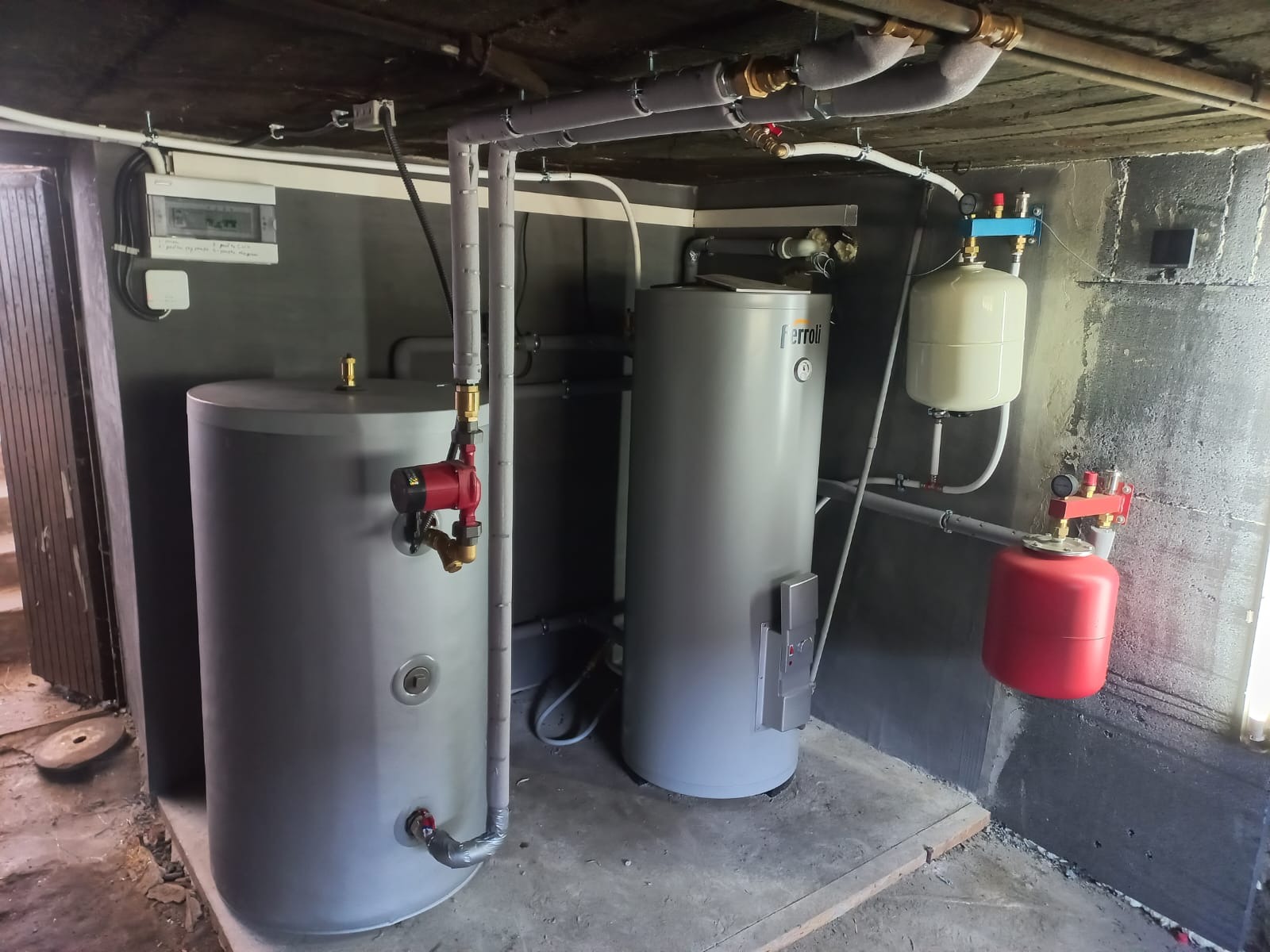 Dwa szare zbiorniki akumulacyjne z czerwonymi pompami obiegowymi oraz czerwonym zbiornikiem przeponowym w kotłowni z szarymi ścianami i rurami instalacyjnymi pod sufitem.