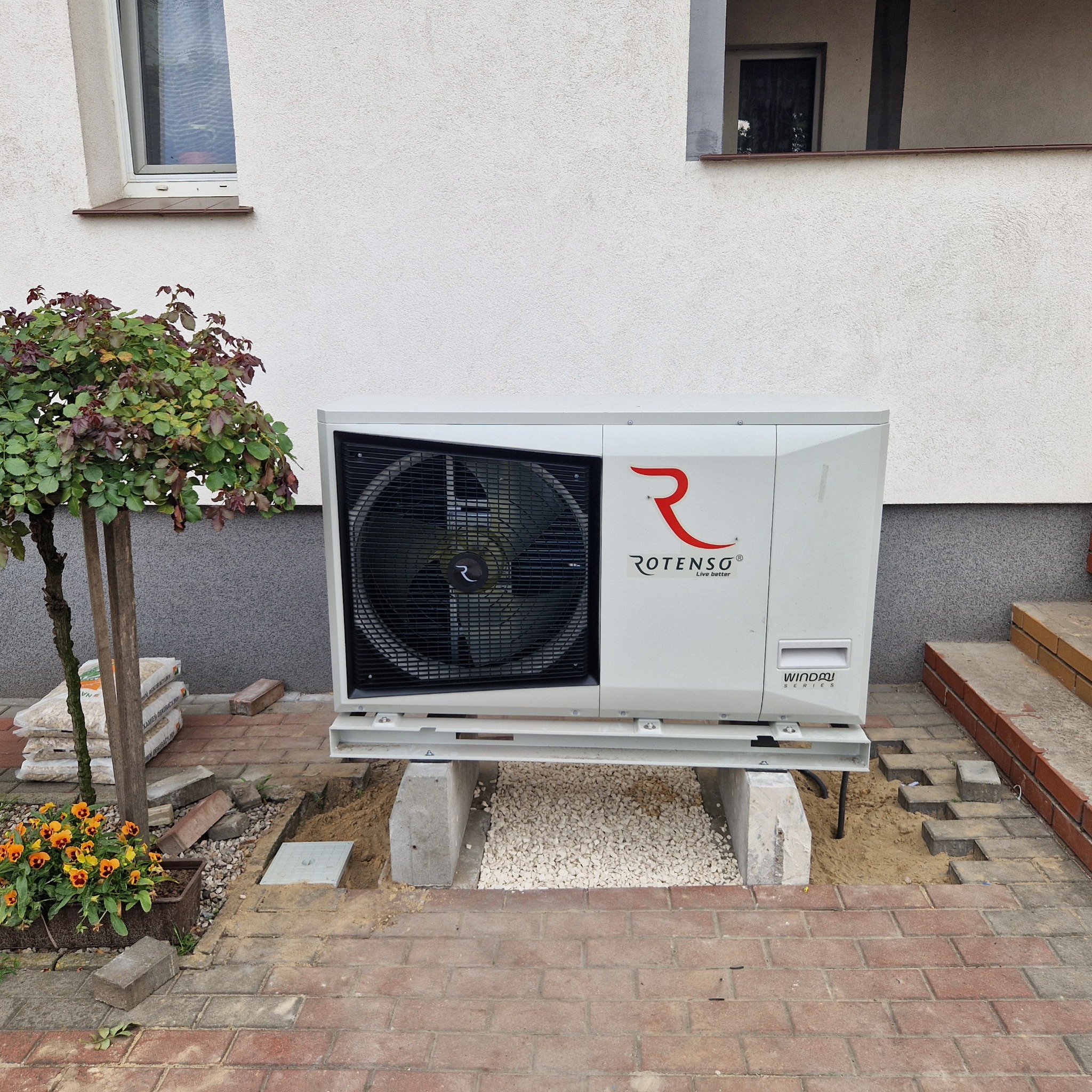Biała pompa ciepła Rotenso Windom Series zainstalowana na betonowych podstawach przed domem, obok klombu z bratkami i worków z granulatem.