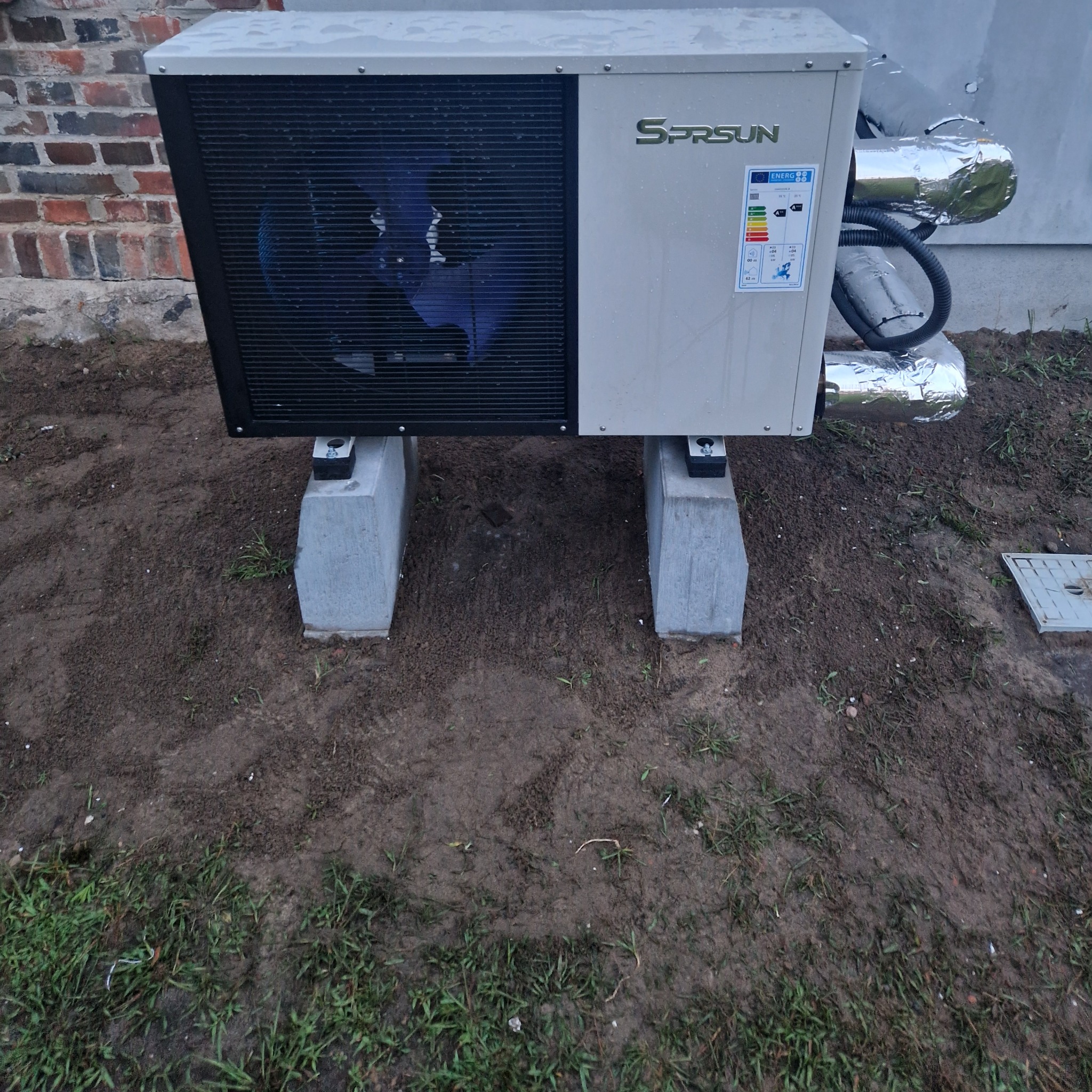 Zewnętrzna jednostka pompy ciepła SPRSUN zamontowana na betonowych wspornikach, z widocznymi przewodami izolowanymi folią aluminiową, na tle ceglanego budynku i trawy.