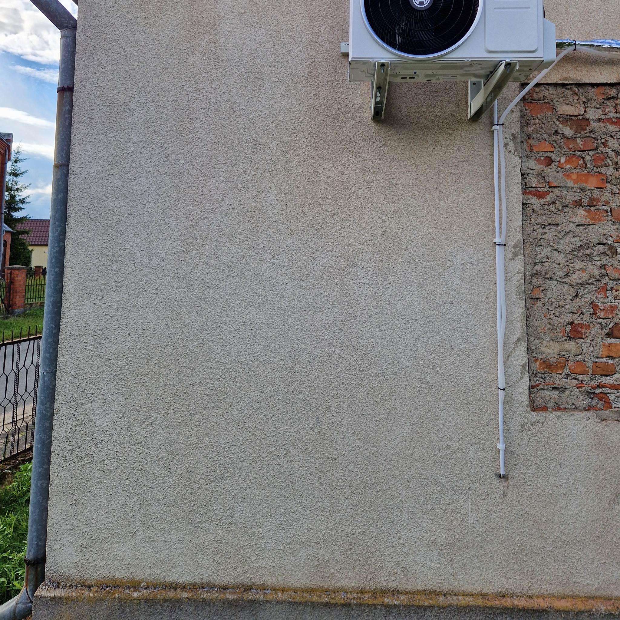 Zewnętrzna jednostka klimatyzacji zamontowana na elewacji budynku z widocznymi przewodami i fragmentem muru z cegły.