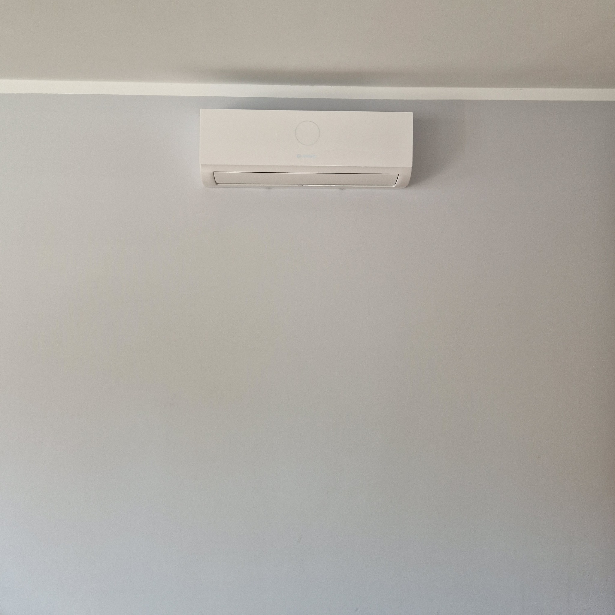 Biały klimatyzator ścienny zamontowany na szarej ścianie z białą listwą podsufitową.