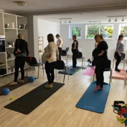 Grupa kobiet w ciąży ćwiczy jogę na matach w jasnym pomieszczeniu z oknami, widoczne piłki do ćwiczeń i butelka wody.