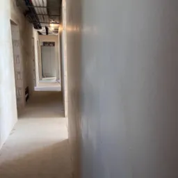 Świeżo położone, jasnoszare ściany w korytarzu budynku w stanie deweloperskim, widoczne instalacje pod sufitem i surowa podłoga.