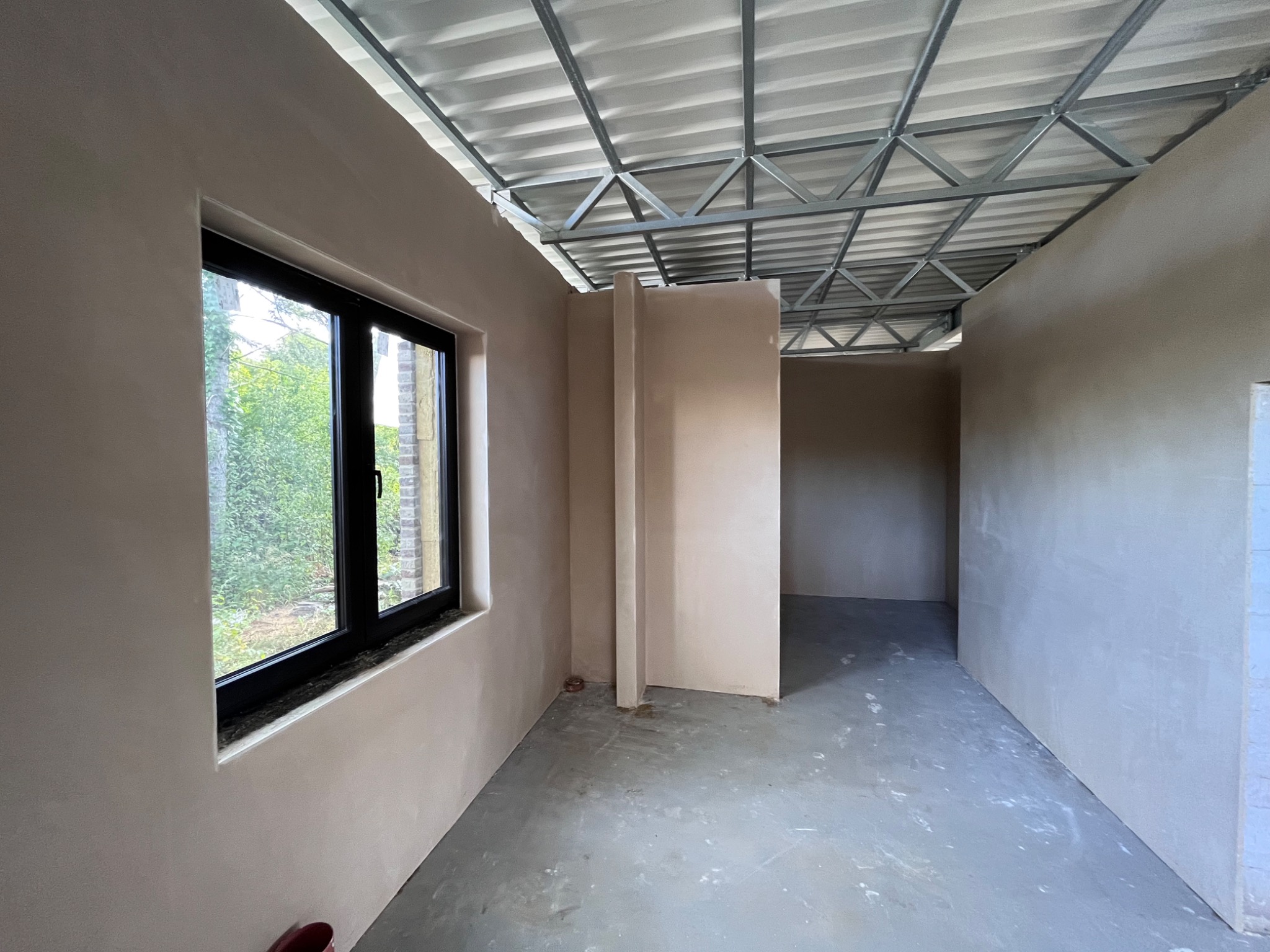 Wnętrze pomieszczenia po nałożeniu jasnych, gładkich tynków na ściany; widoczne czarne ramy okienne i metalowa konstrukcja sufitu.