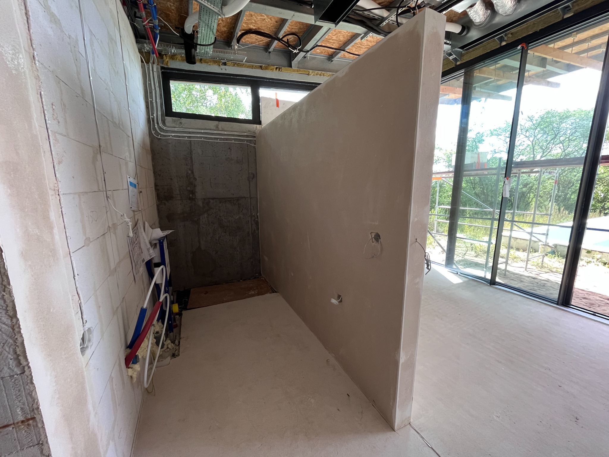 Wnętrze w stanie deweloperskim z wygładzonymi ścianami i podłogą, widoczne instalacje pod sufitem oraz okno z widokiem na zewnątrz.