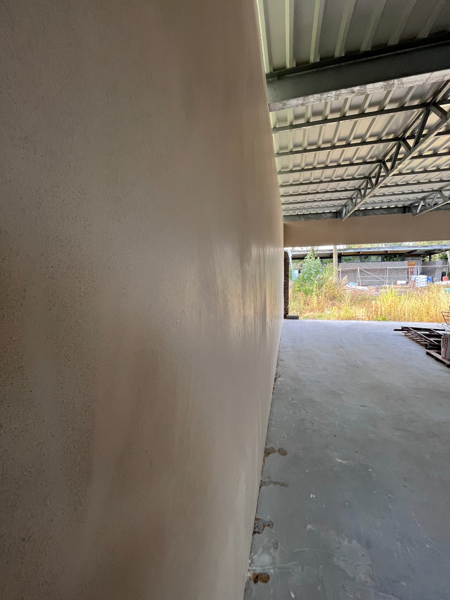 Świeżo nałożony, jasny tynk na ścianie wewnętrznej budynku w surowym stanie, widoczne zacieki na posadzce, metalowa konstrukcja dachu.