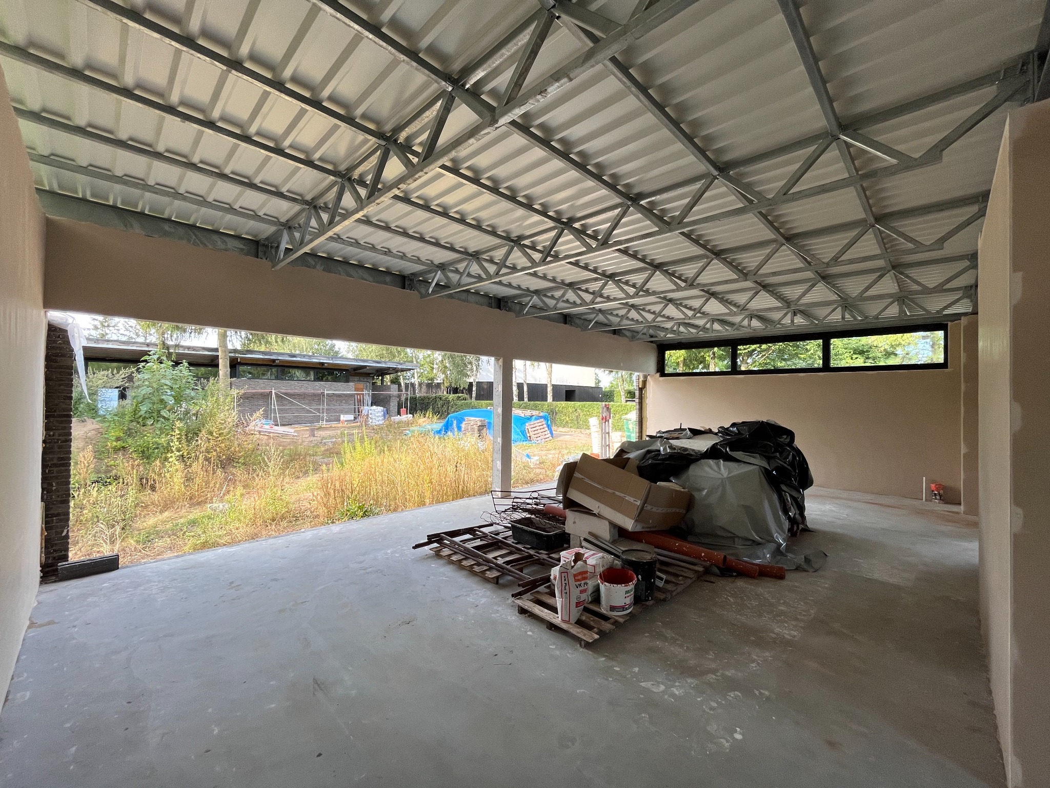 Surowe, otynkowane wnętrze budynku w trakcie prac wykończeniowych; widoczna metalowa konstrukcja dachu, resztki materiałów budowlanych na podłodze i widok na zewnątrz przez otwarty, nieoszklony...