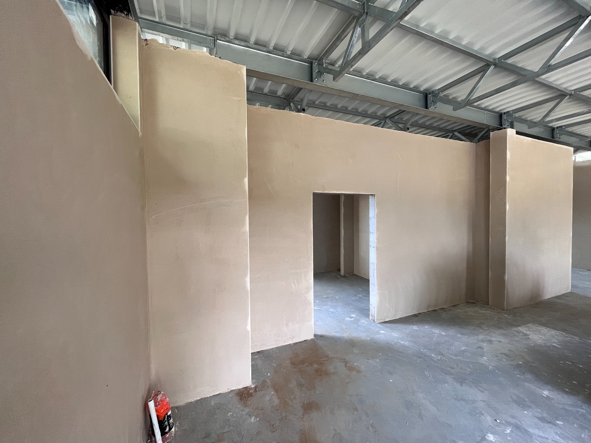 Wnętrze hali w trakcie prac tynkarskich: ściany pokryte świeżym, jasnym tynkiem, widoczna metalowa konstrukcja dachu i fragment worka z zaprawą przy ścianie.
