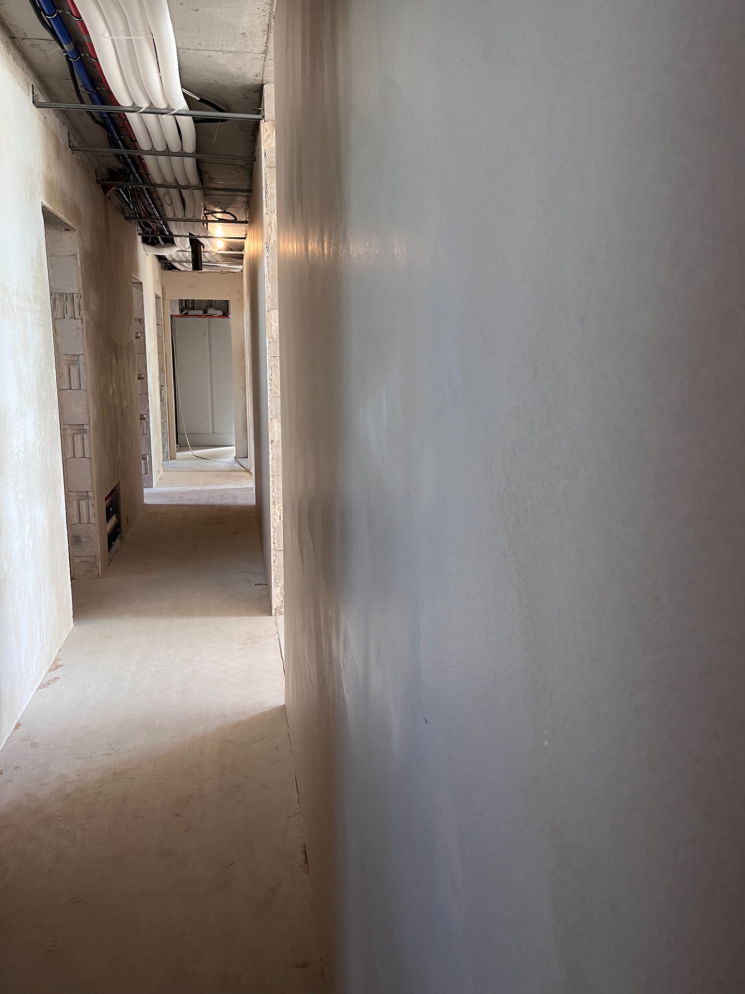 Świeżo położone, jasnoszare ściany w korytarzu budynku w stanie deweloperskim, widoczne instalacje pod sufitem i surowa podłoga.