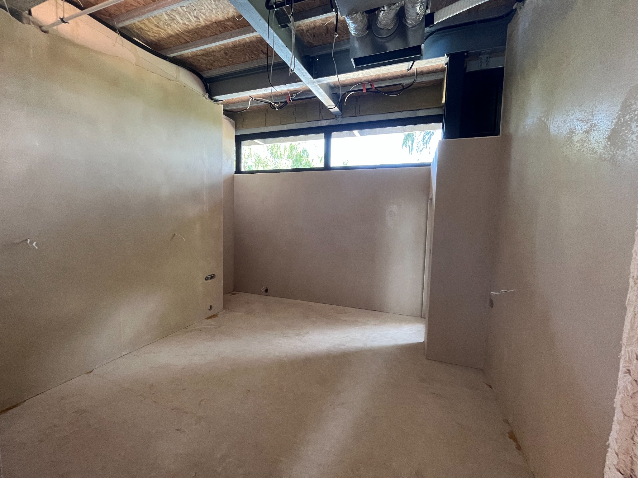 Wnętrze pomieszczenia w trakcie prac wykończeniowych, ściany pokryte świeżo nałożoną, jasną warstwą tynku, widoczne okno z czarną ramą i fragment instalacji pod sufitem.