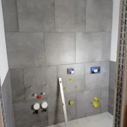 Ściana łazienki w trakcie układania szarych, matowych płytek wielkoformatowych, widoczne otwory na armaturę, poziomica oparta o ścianę, narzędzia glazurnicze na podłodze.