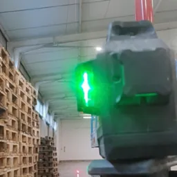 Laser krzyżowy, ustawiony na podnośniku w hali magazynowej, wyznacza pion i poziom. Widoczne palety i oświetlenie sufitowe.