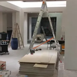 Wnętrze w trakcie remontu: drabina, płyty gipsowe, worki z zaprawą. Widoczne narzędzia i oświetlenie sufitowe. Etap prac wykończeniowych w jasnym pomieszczeniu.