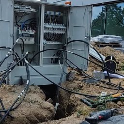 Electro Alpinist Dominik Wójcik - Otwarta skrzynka elektryczna z licznymi kablami na zewnątrz, umieszczona w wykopie na tle nowoczesnego budynku z szarą elewacją i błękitnym niebem.