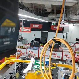 Electro Alpinist Dominik Wójcik - Żółty podnośnik koszowy z przewodami w supermarkecie, prawdopodobnie używany do instalacji oświetlenia lub innych prac na wysokościach w przestrzeni handlowej.