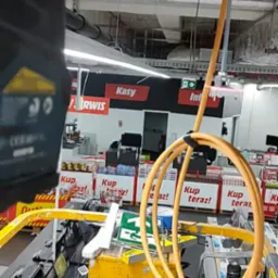 Żółty podnośnik koszowy z przewodami w supermarkecie, prawdopodobnie używany do instalacji oświetlenia lub innych prac na wysokościach w przestrzeni handlowej.