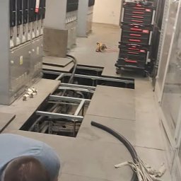 Electro Alpinist Dominik Wójcik - Prace elektryczne w serwerowni: otwarte podłogi techniczne, widoczne kable i instalacje. Elektryk pochylony nad pracą. Rozdzielnice elektryczne w tle.