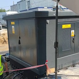 Electro Alpinist Dominik Wójcik - Montaż nowej, szarej stacji transformatorowej. Widoczne kable w ziemi, częściowo ułożona kostka brukowa i pracownik w odblaskowej kamizelce.