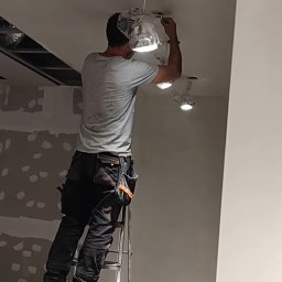 Electro Alpinist Dominik Wójcik - Elektryk na drabinie montuje oświetlenie sufitowe w Pyrzycach. Widoczne kable i oprawy zabezpieczone folią. Ściany przygotowane do malowania.