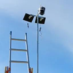 Kamera monitoringu na słupie z panelami solarnymi, widziana z dołu obok drabiny. Błękitne niebo w tle. Drobne kable zwisają ze sprzętu.