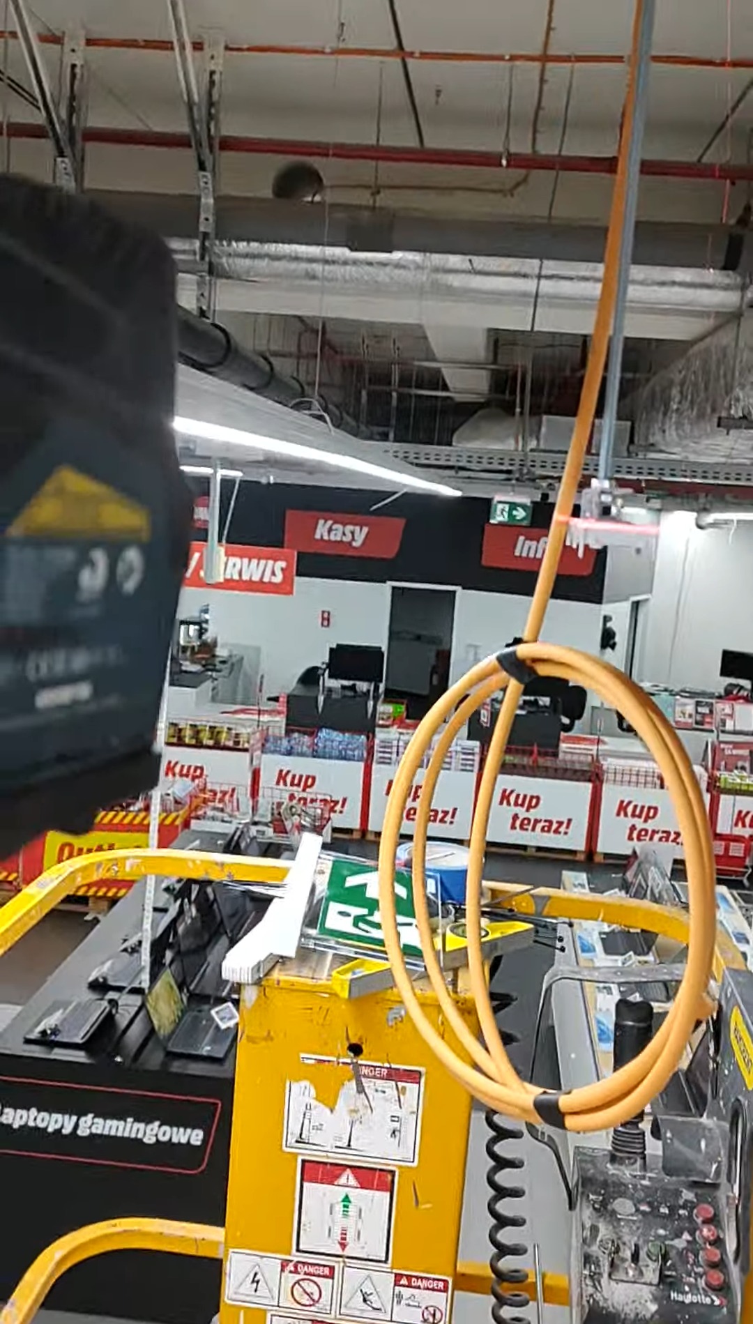 Żółty podnośnik koszowy z przewodami w supermarkecie, prawdopodobnie używany do instalacji oświetlenia lub innych prac na wysokościach w przestrzeni handlowej.