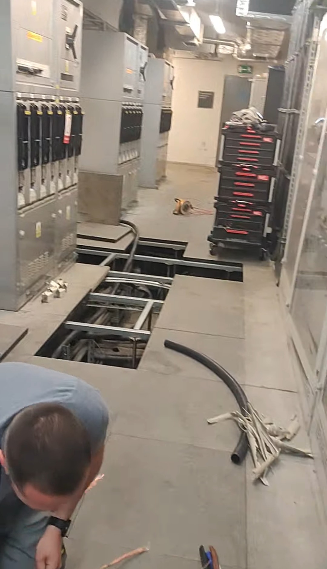 Prace elektryczne w serwerowni: otwarte podłogi techniczne, widoczne kable i instalacje. Elektryk pochylony nad pracą. Rozdzielnice elektryczne w tle.