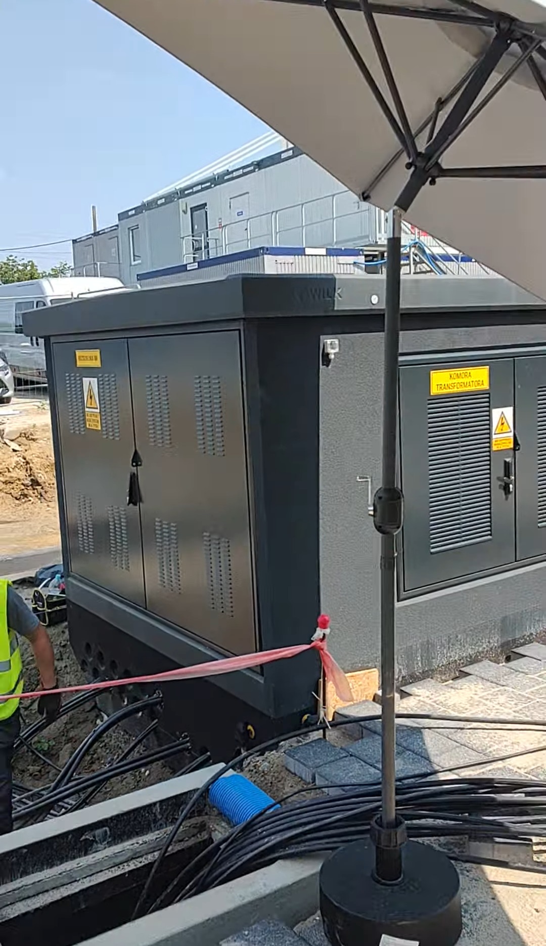 Montaż nowej, szarej stacji transformatorowej. Widoczne kable w ziemi, częściowo ułożona kostka brukowa i pracownik w odblaskowej kamizelce.