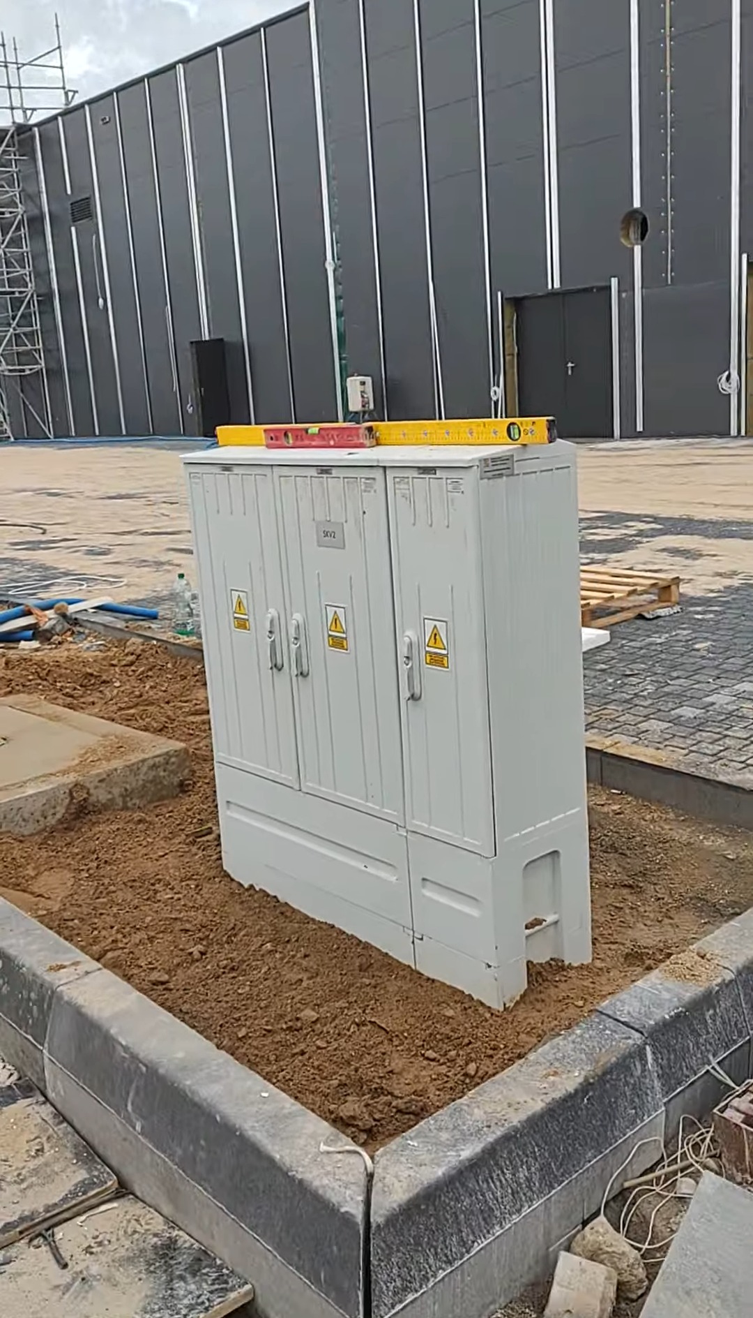 Szara szafa elektryczna z żółtą poziomicą na górze, umieszczona w ziemi obok kostki brukowej i krawężnika, na tle budynku z szarej blachy.