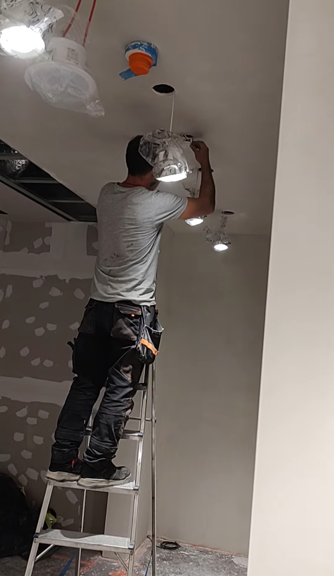 Elektryk na drabinie montuje oświetlenie sufitowe w Pyrzycach. Widoczne kable i oprawy zabezpieczone folią. Ściany przygotowane do malowania.