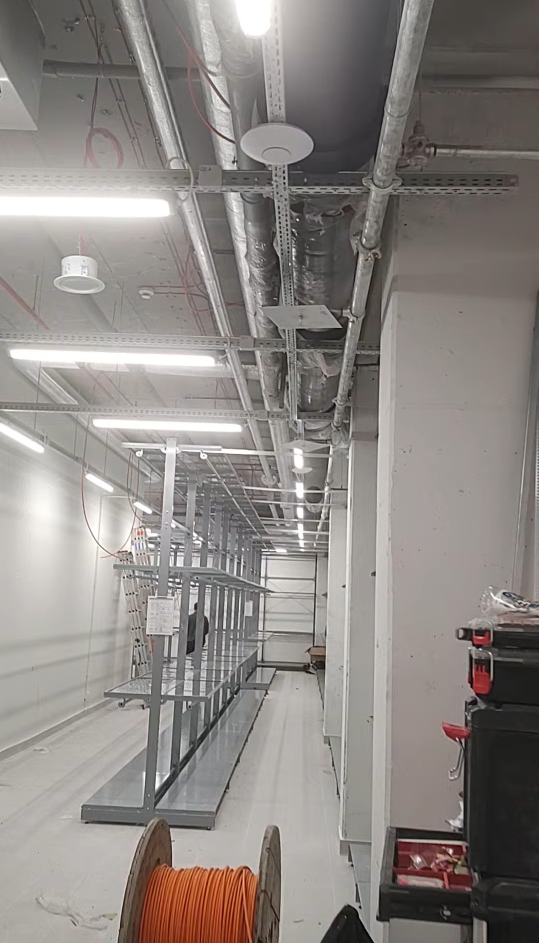 Wnętrze hali w trakcie instalacji elektrycznej: widoczne kable na szpuli, metalowe koryta kablowe pod sufitem i oświetlenie. Częściowo widoczny regał i skrzynka narzędziowa.