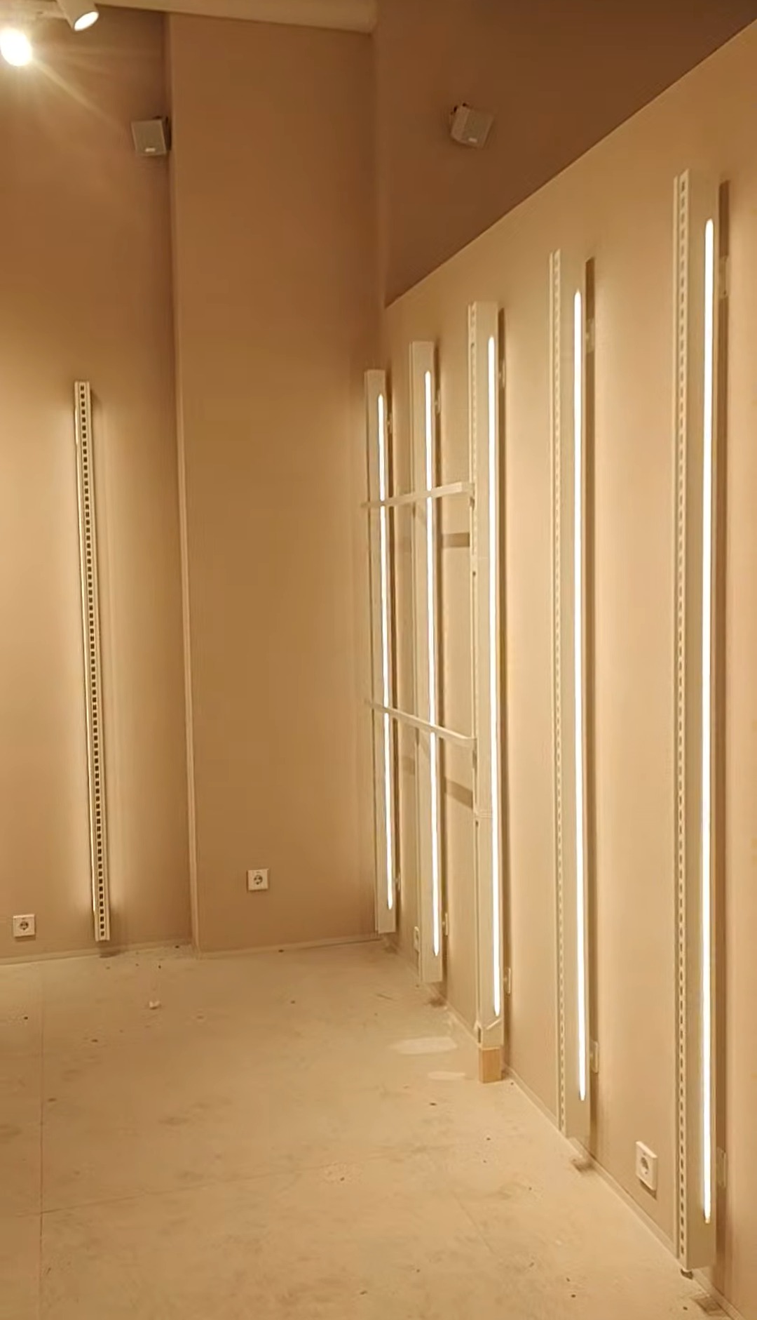 Zamontowane pionowe listwy oświetleniowe LED na ścianie w beżowym pomieszczeniu. Widoczne gniazdka elektryczne. Oświetlenie w stylu industrialnym.
