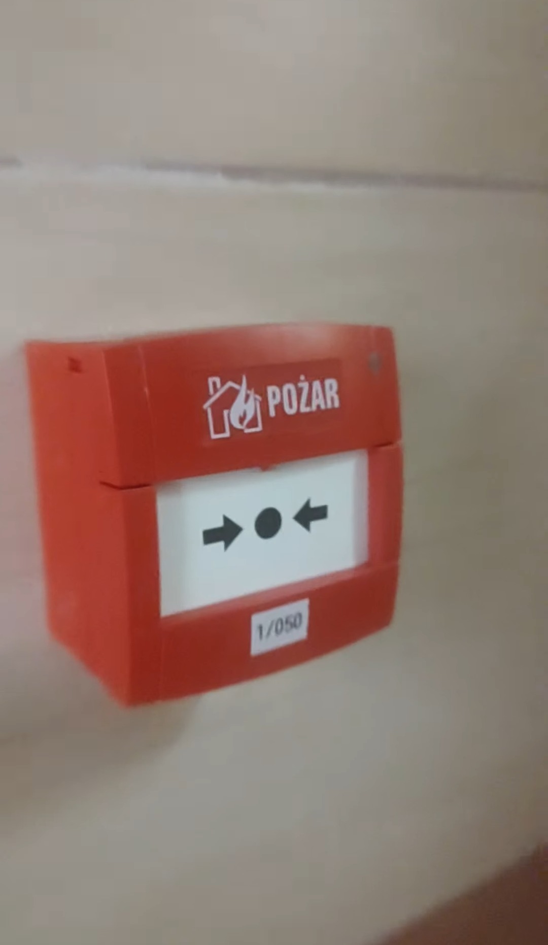 Czerwony, natynkowy ręczny ostrzegacz pożarowy z symbolem płomienia i napisem 'POŻAR', zamontowany na ścianie. Widoczny numer seryjny 1/050.