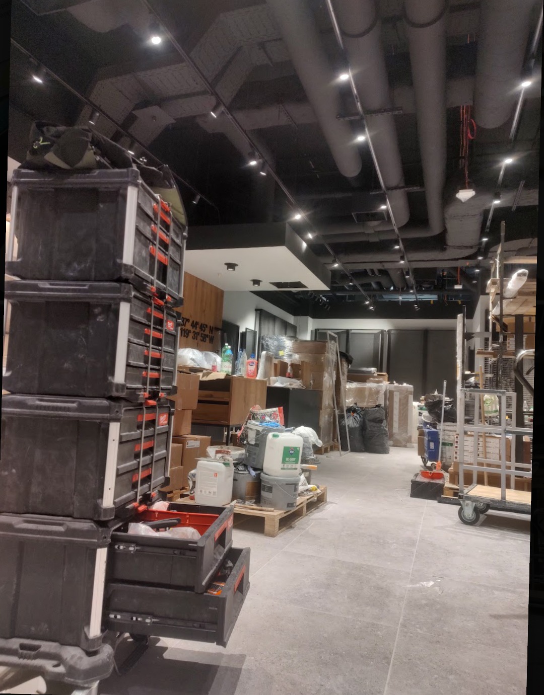 Wnętrze sklepu w trakcie prac instalacyjnych: skrzynie narzędziowe na pierwszym planie, w tle materiały budowlane, widoczne instalacje pod sufitem i oświetlenie.