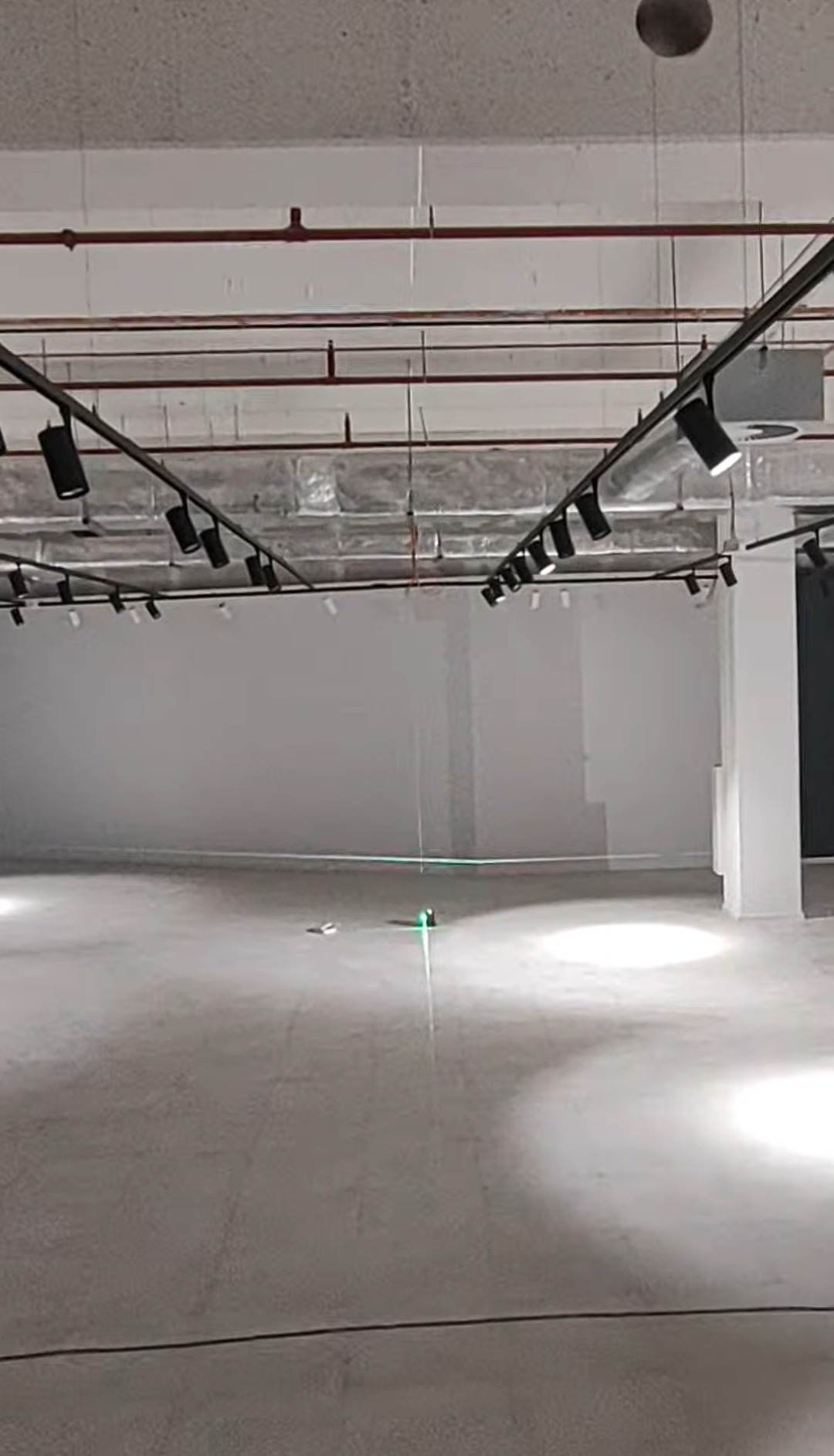 Wnętrze komercyjne z surowym betonem, oświetleniem szynowym i laserowym niwelatorem wskazującym poziom. Widoczne instalacje pod sufitem.