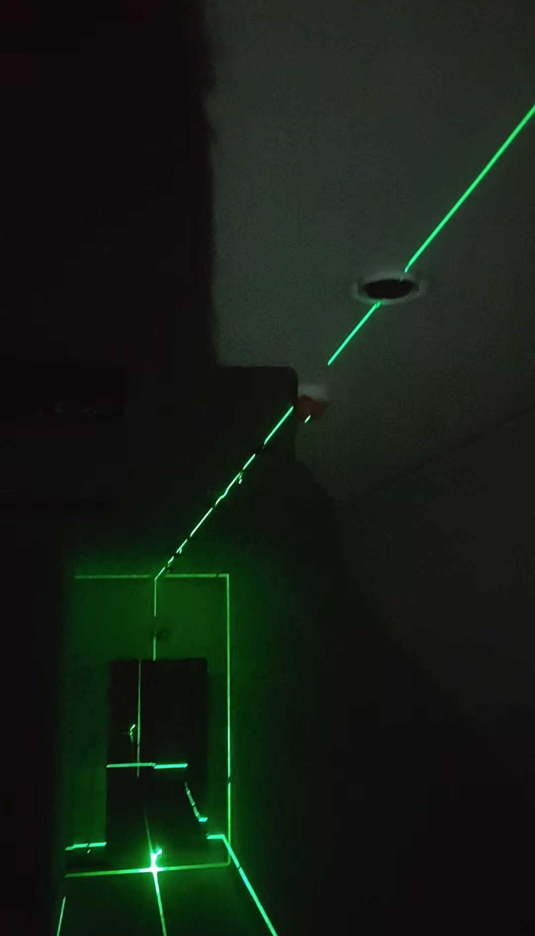Zielony laser krzyżowy w ciemnym pomieszczeniu, wyznaczający linie na ścianach i suficie, prawdopodobnie podczas prac instalacyjnych lub pomiarowych.