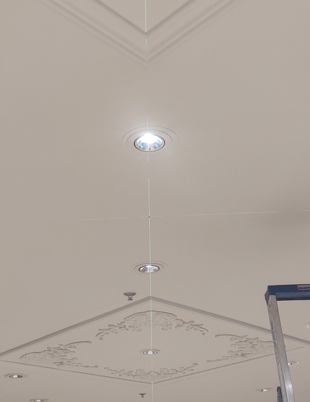 Biały sufit z dekoracyjną sztukaterią i oświetleniem punktowym, wyznaczonym laserowo, z fragmentem drabiny malarskiej w prawym dolnym rogu.