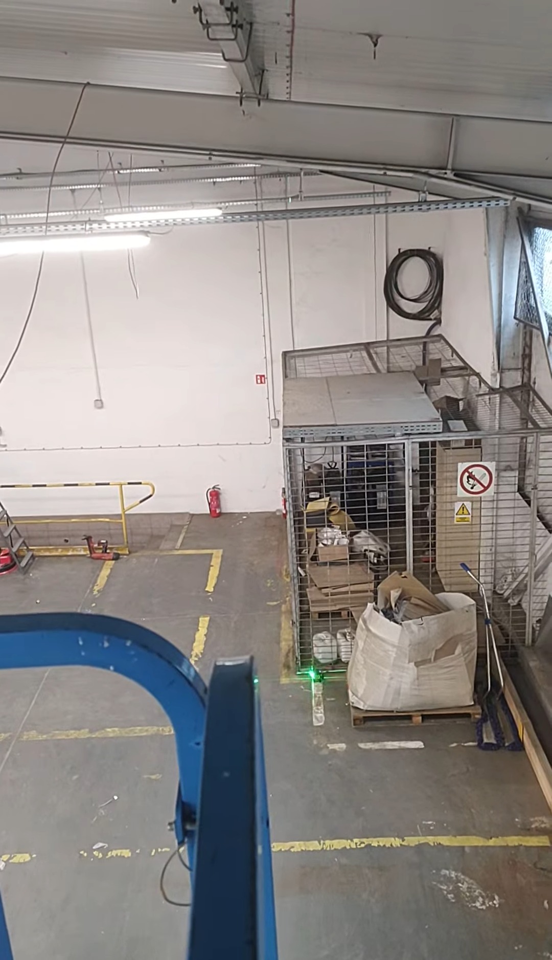 Widok z góry na magazyn z instalacjami pod sufitem i klatką z zabezpieczeniami. Widoczny wózek podnośnikowy z poziomnicą laserową.