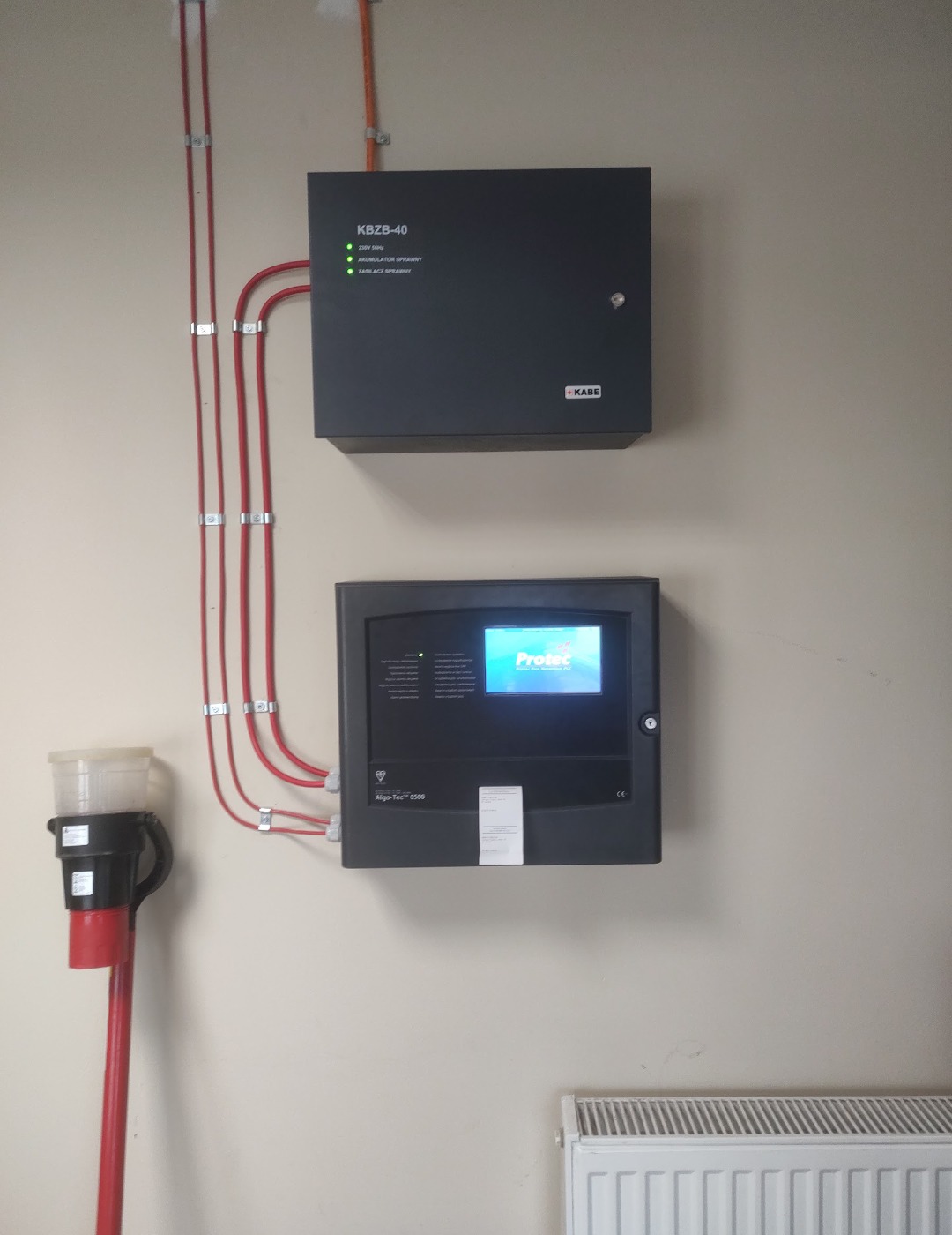 Zamontowana centrala alarmowa Protec Algo-Tec 5500 z sygnalizatorem i okablowaniem na ścianie. Widoczne diody i ekran. Dolny kadr z grzejnikiem.