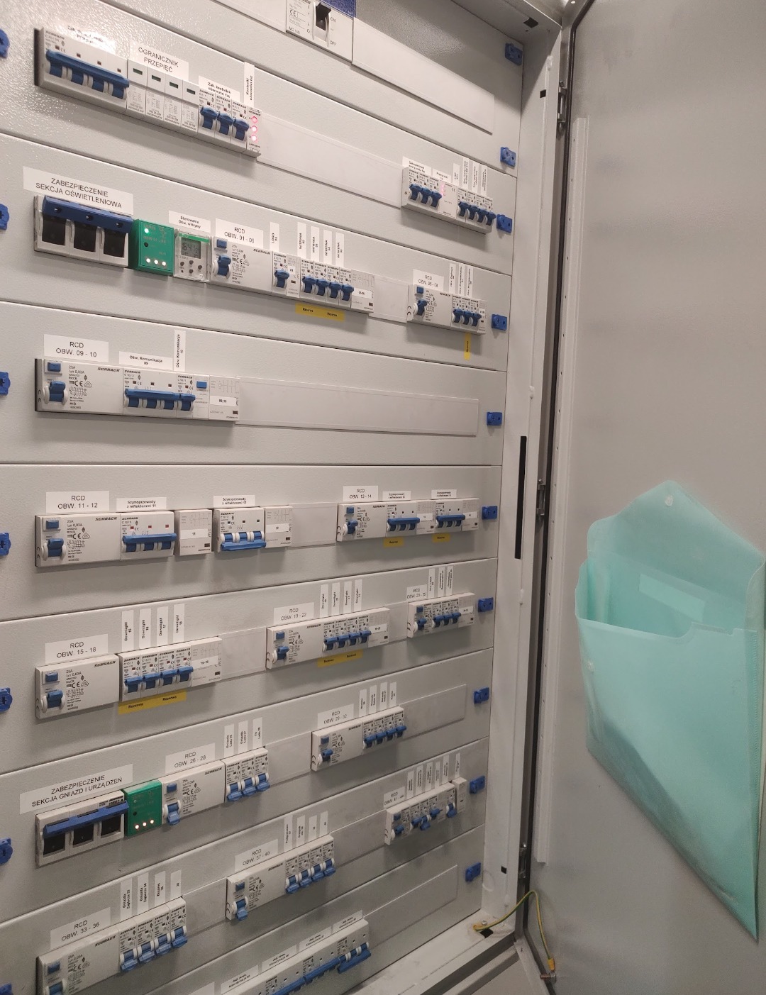 Otwarta szafa elektryczna z licznymi zabezpieczeniami różnicowoprądowymi (RCD) i wyłącznikami, widoczne opisy obwodów. Wewnątrz dokumentacja w kieszeni.