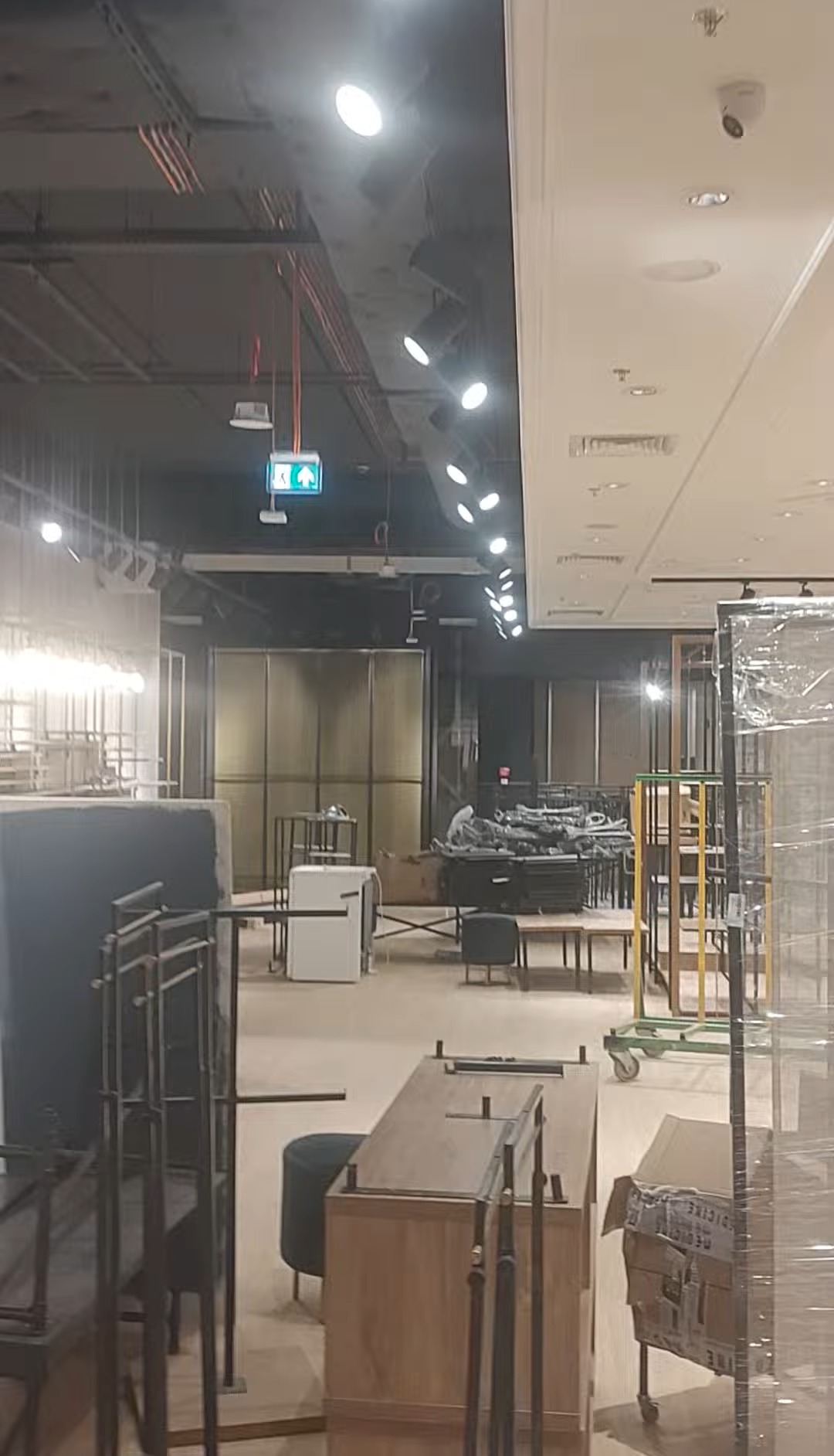 Wnętrze sklepu w trakcie instalacji oświetlenia, widoczne lampy sufitowe i kamery. Elementy wyposażenia w tle, surowy stan przed otwarciem.