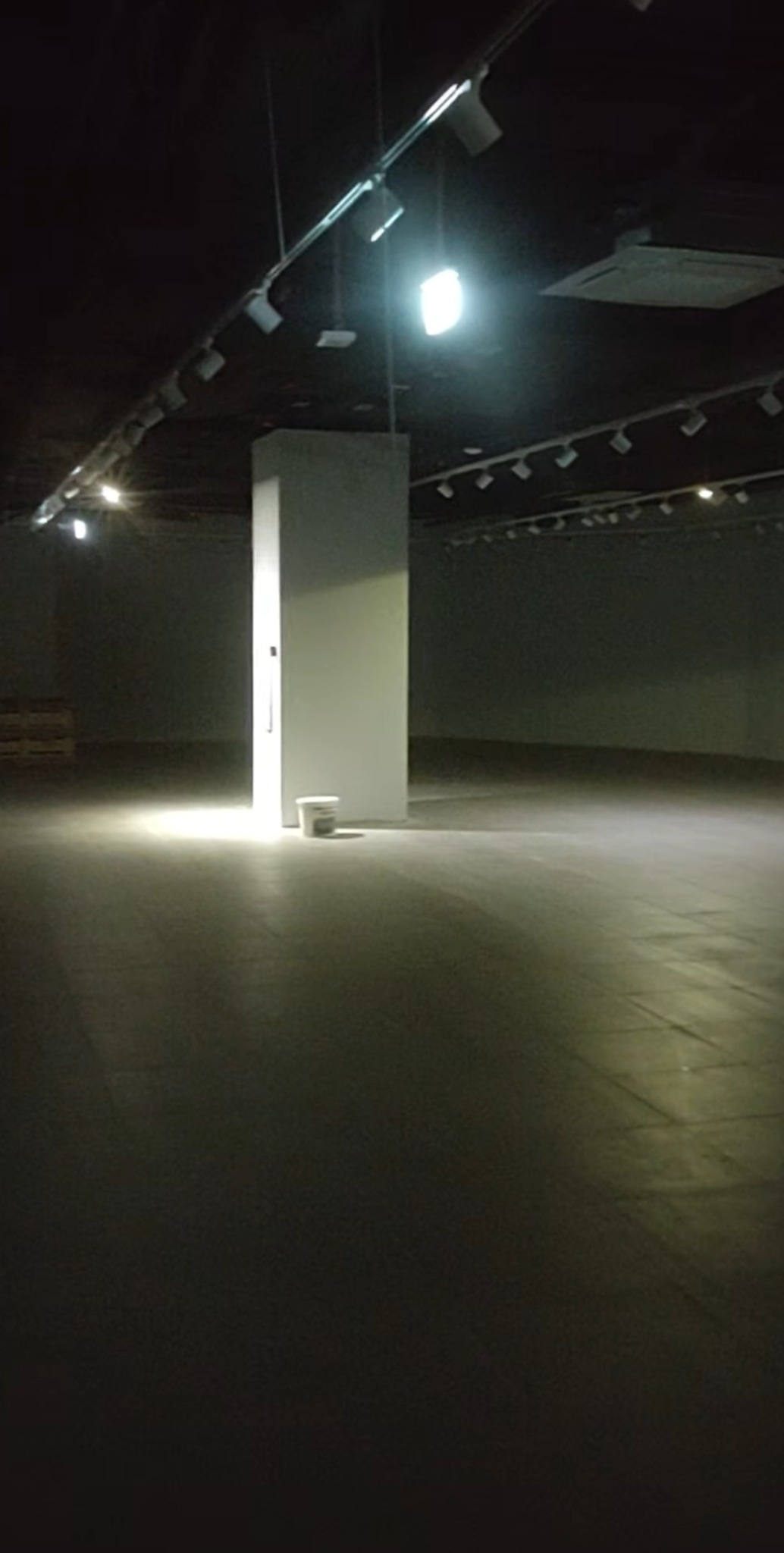 Pusta hala z betonową podłogą i oświetleniem sufitowym, biały filar i wiadro z farbą. Przygotowanie do malowania, remont w trakcie.