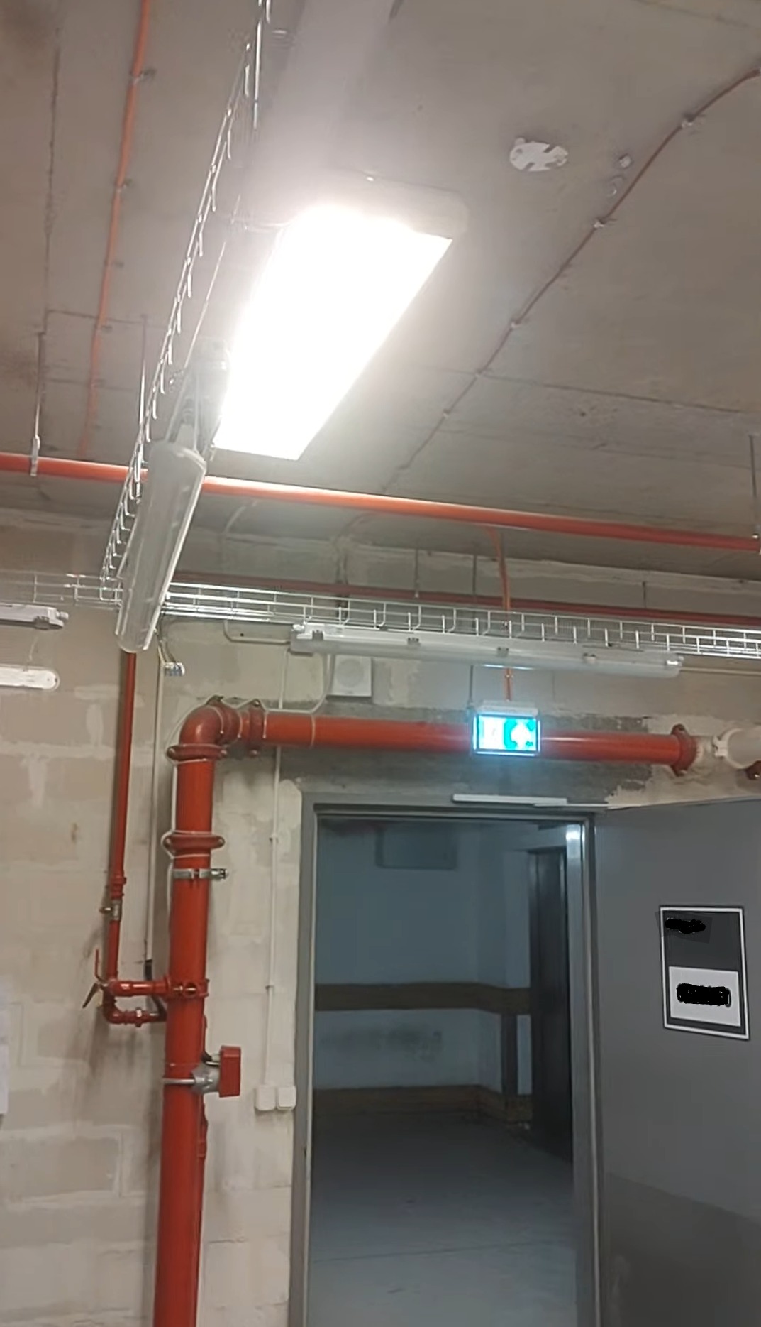 Instalacja oświetleniowa w hali z widocznymi przewodami w korytkach, rurami ppoż. i znakiem ewakuacyjnym nad drzwiami. Surowy betonowy strop.