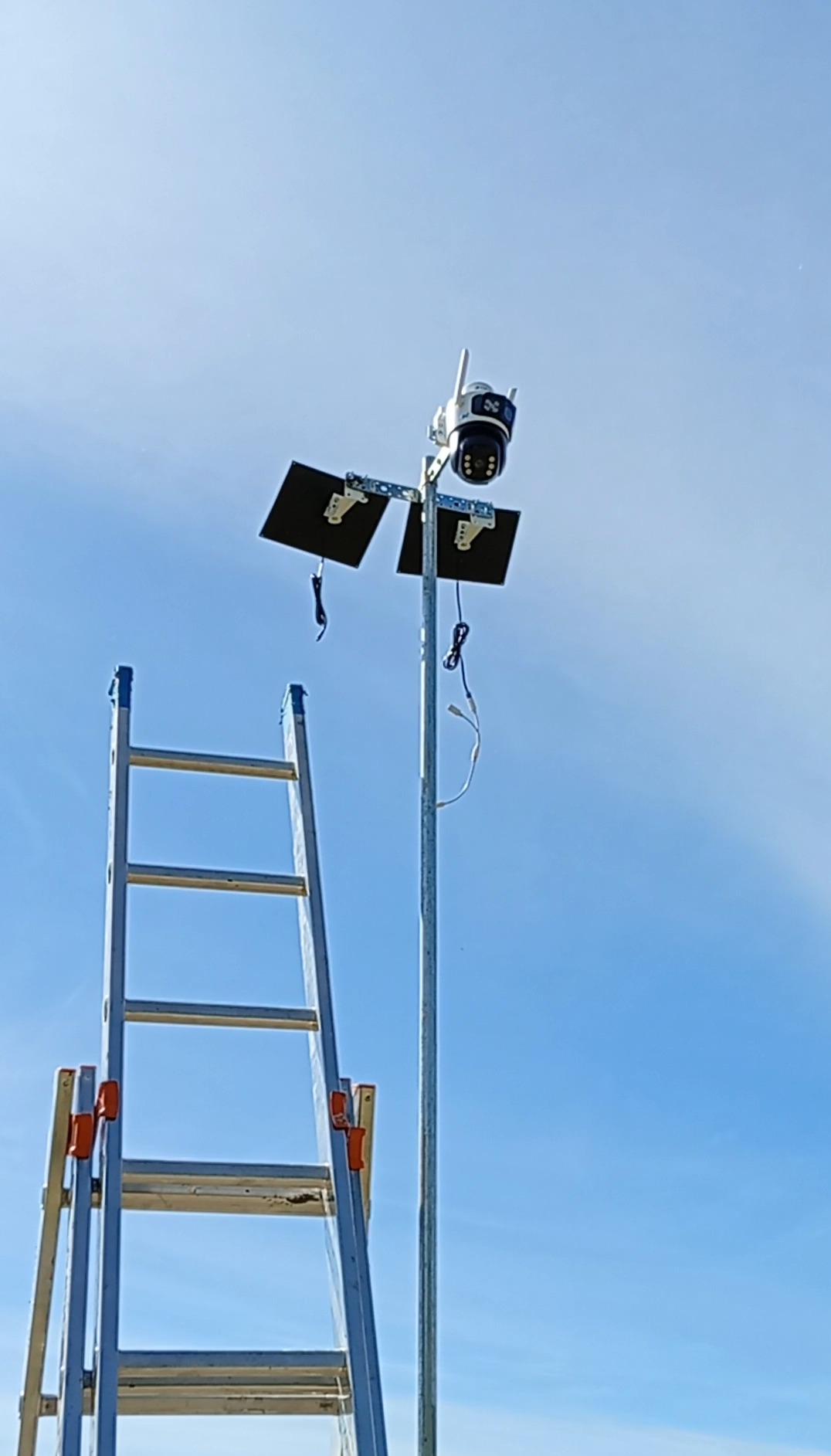 Kamera monitoringu na słupie z panelami solarnymi, widziana z dołu obok drabiny. Błękitne niebo w tle. Drobne kable zwisają ze sprzętu.